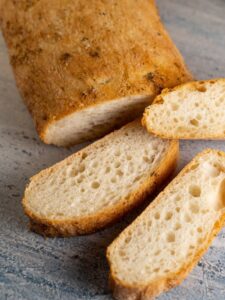 A close-up of a freshly baked artisan bread loaf with several slices on a textured surface.